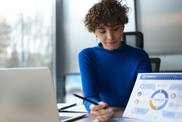 Uma pessoa de cabelos curtos e cacheados, vestindo um suéter azul, senta-se em uma mesa com um laptop e aponta para um gráfico de relatório financeiro com uma caneta, demonstrando como fazer modelo de gestão em um ambiente de escritório.