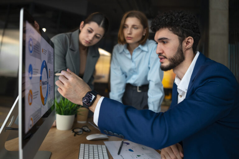 Três profissionais se reúnem em torno de um monitor de computador que exibe tabelas e gráficos de casas. Um homem de terno azul aponta para a tela enquanto duas mulheres observam atentamente. Uma planta e papéis estão sobre a mesa.