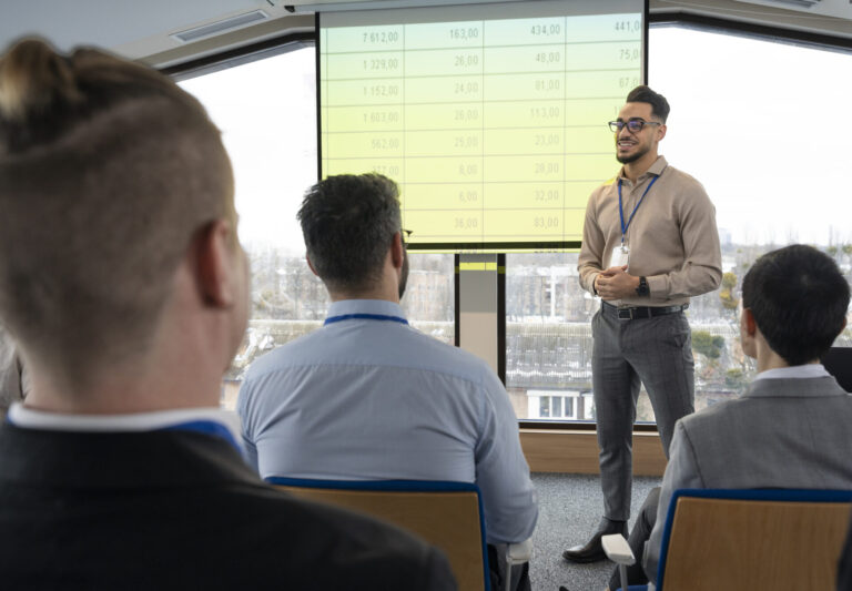 Um homem em trajes de negócios está diante de um grupo, apresentando-se ao lado de uma tela que exibe uma planilha sobre formação de equipes. O público ouve atentamente em um ambiente de escritório moderno e iluminado que lembra casas de trabalho inovadoras.