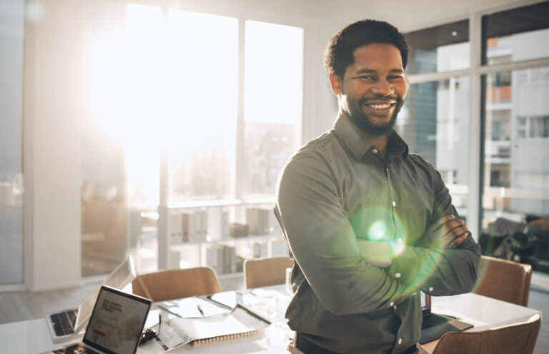 Um homem sorridente com os braços cruzados está em um escritório iluminado pelo sol, ao lado de uma mesa com um laptop, papéis e óculos, representando a missão visão valores. A luz do sol atravessa grandes janelas ao fundo.