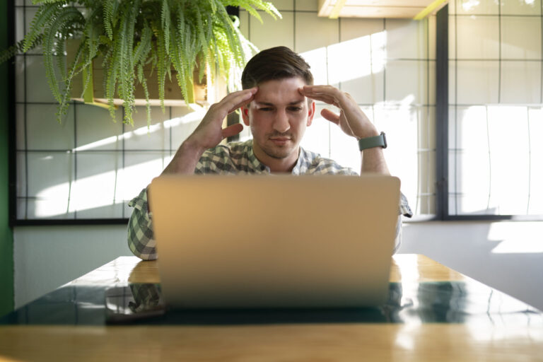 Um homem está sentado em uma mesa com um laptop, segurando as têmporas e parecendo estressado ou frustrado, provavelmente devido à falta de software lotérica. Um smartphone está sobre a mesa e um vaso de plantas está pendurado ao fundo.