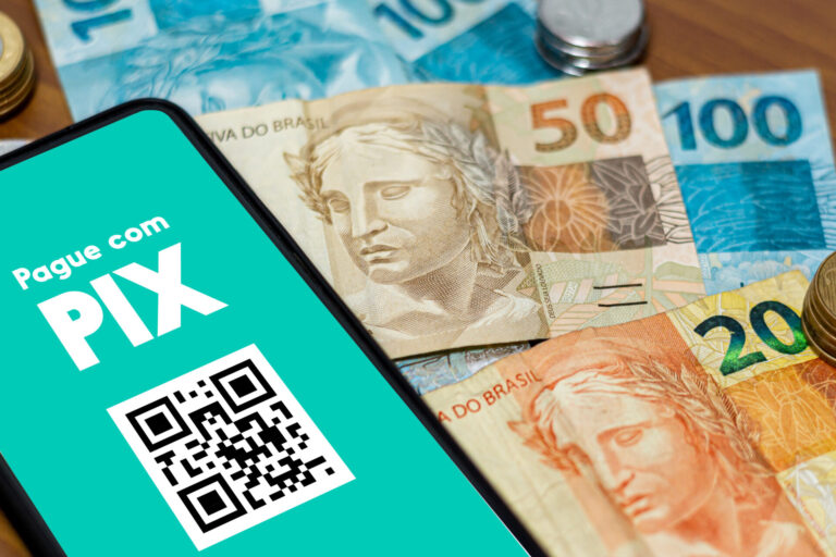 A tela de um smartphone mostra o Pague com PIX em um aplicativo e um código QR, enquanto o dinheiro brasileiro - cédulas e moedas reais - está espalhado ao fundo.