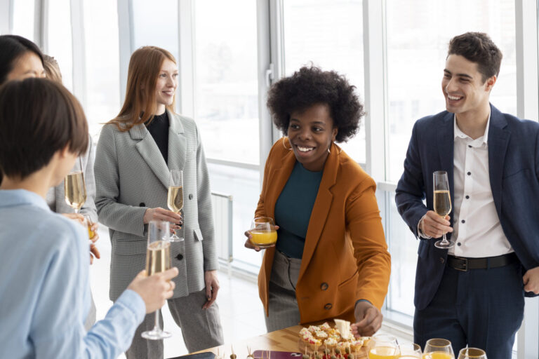 Um grupo de pessoas vestidas profissionalmente desfruta de confraternizações para o tempo lotérico, sorrindo e conversando ao redor de uma mesa com bebidas e lanches em um escritório bem iluminado. Uma pessoa pega a comida enquanto segura um copo de suco de laranja.