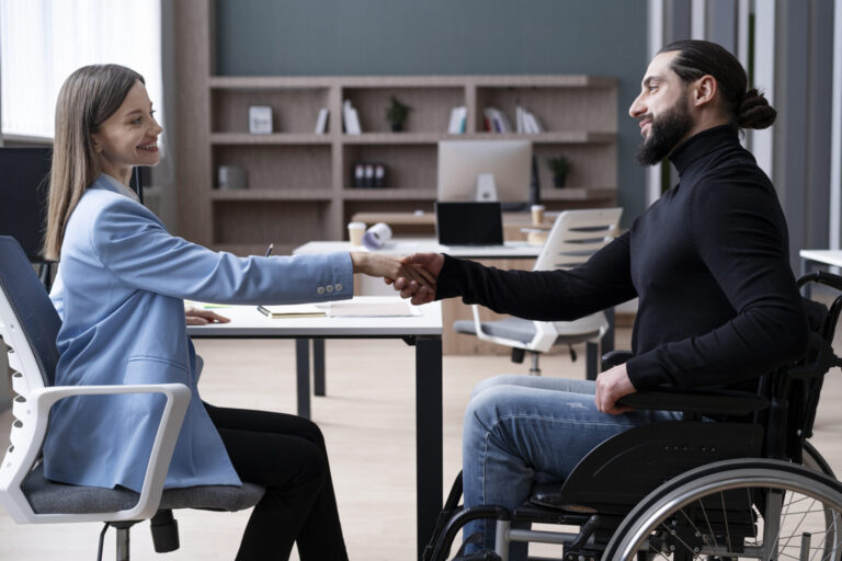 Uma mulher de blazer azul e um homem em uma cadeira de rodas estão sentados um em frente ao outro em uma mesa, sorrindo e apertando as mãos enquanto conversam sobre a caixa preferencial lotérica em um escritório.
