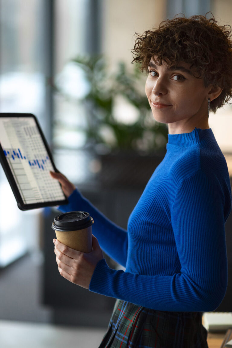 Uma pessoa de cabelos curtos e cacheados, vestindo um suéter azul brilhante, segura uma xícara de café e um tablet que exibe tabelas ou gráficos sobre Notion: como ele pode facilitar sua vida. Eles estão em pé em uma sala e olham por cima do ombro com um leve sorriso.
