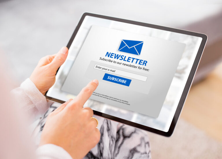 Uma pessoa segurando um tablet com um formulário de assinatura do boletim informativo para sua casa lotérica na tela, com um ícone de envelope azul, um campo de entrada de e-mail e um botão laranja Assinar.