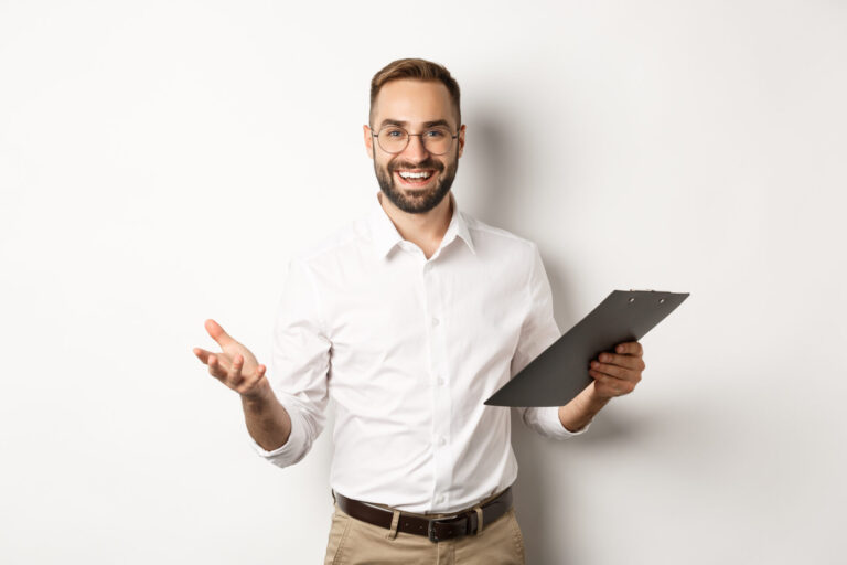 Um homem de óculos e barba, vestindo camisa branca e calça cáqui, está em pé em um fundo branco, sorrindo e segurando uma prancheta em uma das mãos enquanto gesticula sobre operador de caixa e as oportunidades: como crescer na carreira.