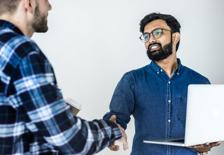 Dois homens estão se cumprimentando com um aperto de mão. Usando técnicas de relacionamento com o cliente, um homem de óculos e camisa azul segura um laptop aberto, enquanto o outro, de camisa xadrez, tem uma xícara de café. Eles parecem estar conversando.