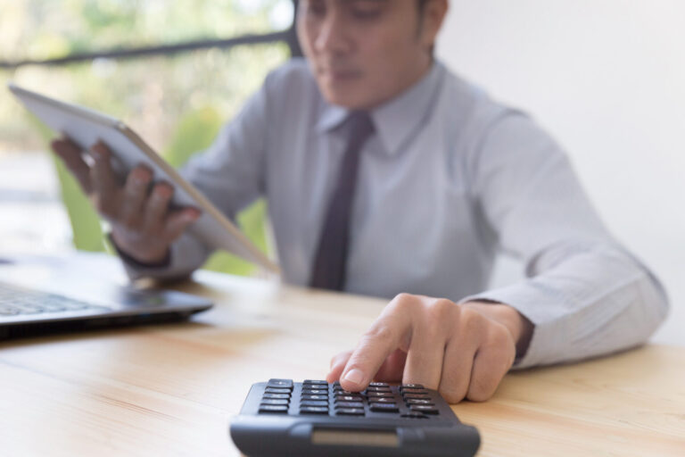 Um homem em traje de negócios está sentado em uma mesa, usando uma calculadora com uma mão e segurando um tablet com a outra, concentrado em calcular comissões funcionários. Um laptop está aberto nas proximidades e o fundo está suavemente desfocado.