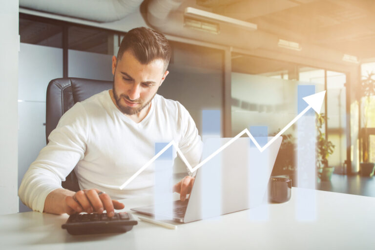 Um homem de camisa branca trabalha em uma mesa com um laptop e uma calculadora. Um gráfico de barras e linhas ascendentes é sobreposto, simbolizando como os serviços podem aumentar a lucratividade e impulsionar o sucesso nos negócios.