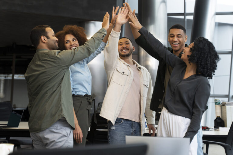 Cinco colegas de trabalho em um escritório, sorrindo e dando um high-five em grupo, comemorando a importância do trabalho em equipe e o sucesso compartilhado. Mesas de escritório, cadeiras e prateleiras são visíveis ao fundo.