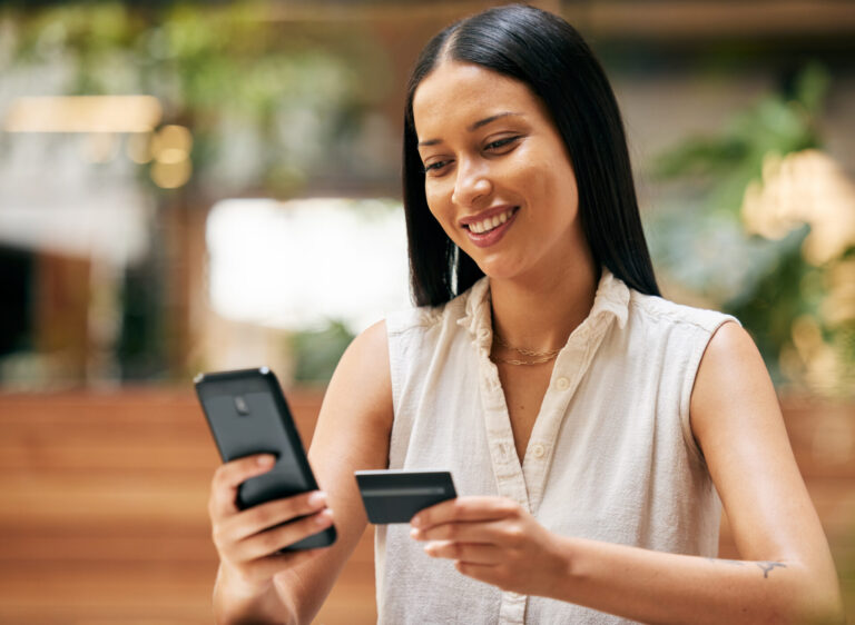 Uma mulher sorridente, com longos cabelos escuros, segura um smartphone em uma mão e um cartão de crédito na outra, parecendo usar as melhores plataformas de pagamento. Ela usa uma blusa branca sem mangas e está sentada em um ambiente interno com vegetação desfocada ao fundo.