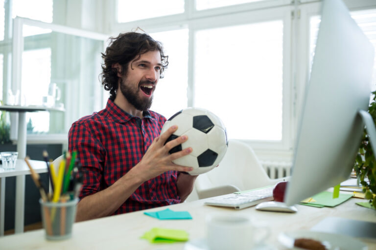 Um homem sorridente com cabelos cacheados e barba segura uma bola de futebol enquanto está sentado em sua mesa com um computador, comemorando enquanto explora lotéricas: melhores sites de apostas esportivas em um ambiente de escritório bem iluminado.