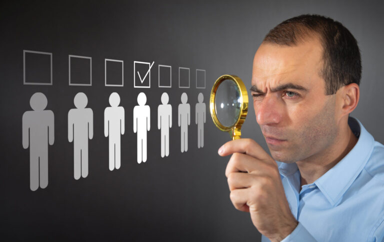 Um homem segurando uma lupa examina uma fileira de ícones de bonecos de palito em um quadro negro. Uma figura está destacada com uma marca de verificação acima dela, representando como identificar o perfil do cliente ideal.