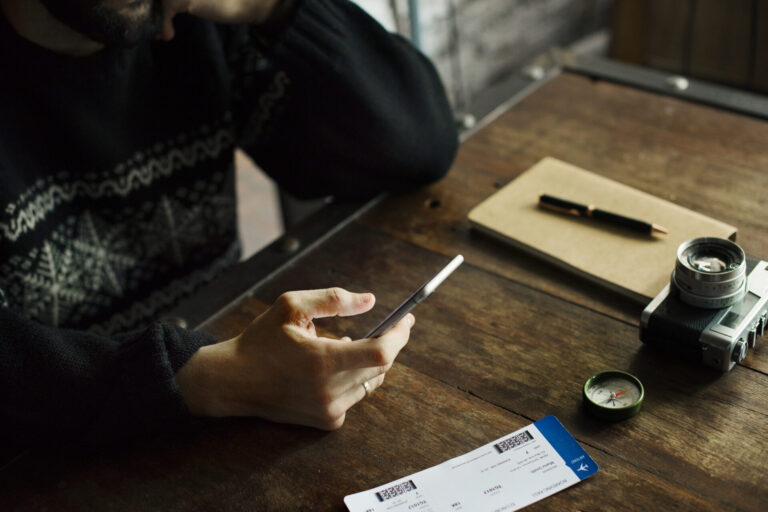 Uma pessoa está sentada em uma mesa de madeira, segurando um smartphone. Sobre a mesa há um caderno com uma caneta, uma câmera, planos de assinatura, uma bússola e dois cartões de embarque de companhias aéreas. A cena sugere o planejamento de uma viagem.