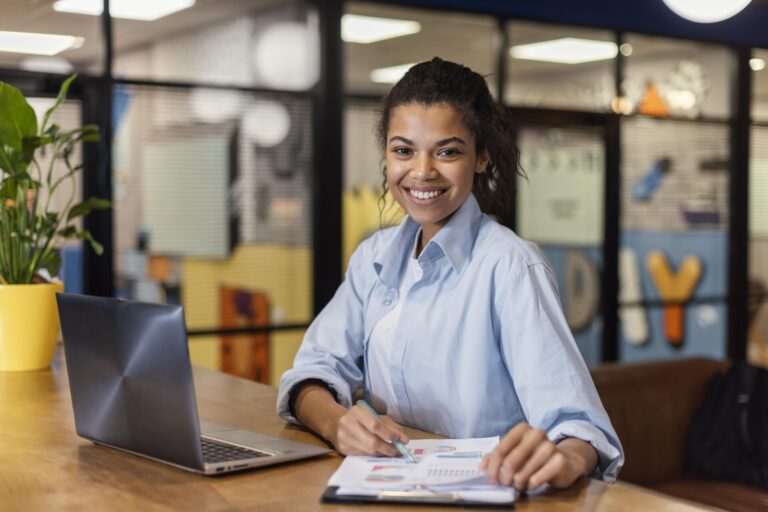 Uma mulher sorridente está sentada em uma mesa com um laptop e um caderno aberto, segurando uma caneta. Vestida com uma camisa azul clara, ela parece estar trabalhando como operadora de caixa lotérica em um escritório moderno e bem iluminado.