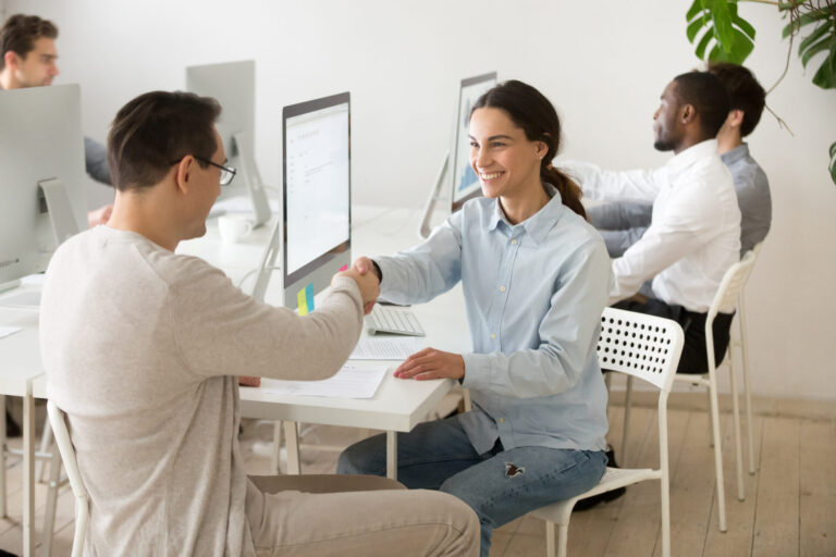Dois colegas sentados em uma mesa em um escritório, sorrindo e apertando as mãos - demonstrando o recrutamento inteligente de colaboradores em lotérica: como criar um ambiente colaborativo e profissional entre a equipe.