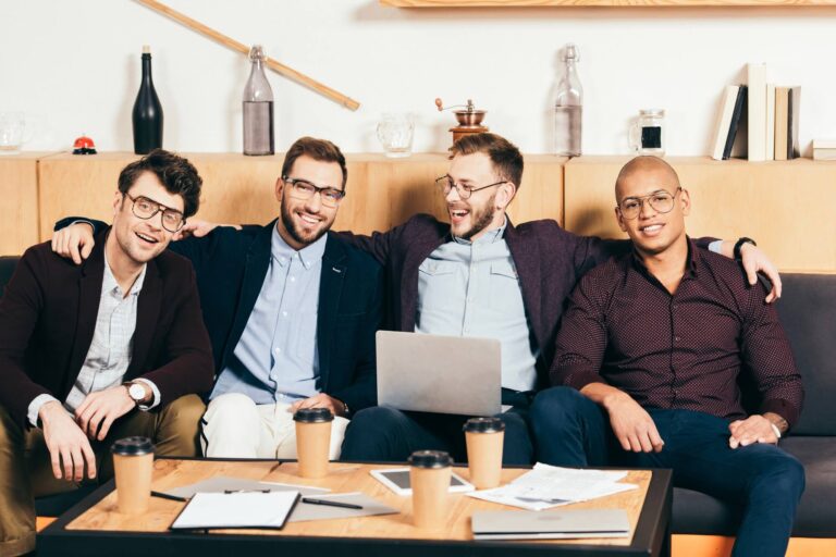 Quatro homens sentados juntos em um sofá, sorrindo e rindo, com xícaras de café, papéis e um laptop - talvez discutindo o software mais recente para lotéricas - em um ambiente de escritório moderno.